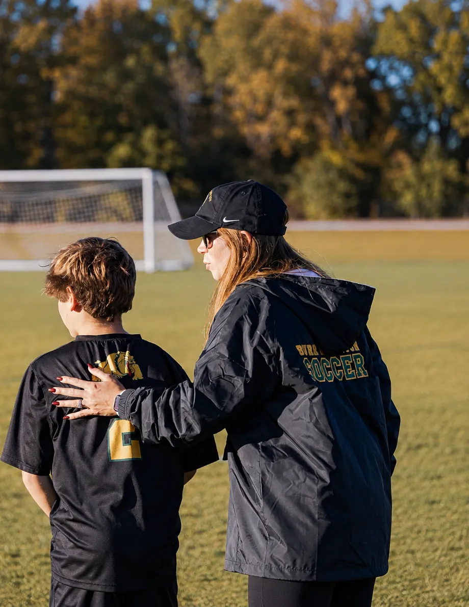 Morgan LaDuca on the sideline in a team soccer jacket and cap, one hand on a young player's shoulder as the two of them watch the field